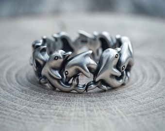 May include: A silver-toned ring featuring a continuous band of sculpted dolphins linked together in a circular formation. The metal has a matte, brushed finish with dark recessed areas that define the eyes and body contours of each dolphin.