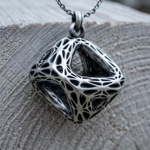 Puede incluir: Un colgante de tono plateado con forma de cubo tridimensional hueco y un intrincado patrón de celosía similar a una red. El colgante cuelga de una cadena plateada a juego, mostrando un acabado oscuro y oxidado dentro de los huecos.