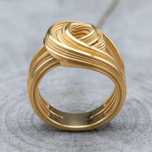 May include: A gold-toned ring featuring a multi-layered, coiled wire design that forms a central knot. The surface of the ring displays a series of parallel, raised lines that wrap around the band, creating a textured, dimensional appearance.