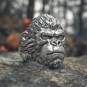 May include: A detailed silver ring shaped like a gorilla's head. The ring features intricate textures and a realistic depiction of the primate's facial features. The ring is set on a textured, gray surface.