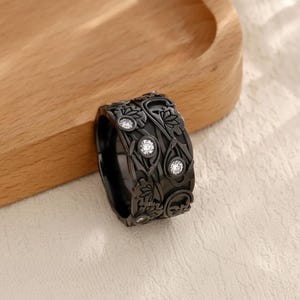May include: A black ring featuring an elaborate floral pattern and clear gemstones. The ring is displayed on a light surface and a wooden tray, highlighting its detailed design and sophisticated appearance.