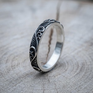 Puede incluir: Un anillo de plata con un patrón en relieve detallado de volutas orgánicas y motivos en forma de hoja. El metal tiene un acabado pulido en la banda interior y un acabado oxidado oscuro en las áreas hundidas del diseño del anillo.