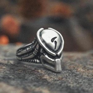 May include: A silver-colored ring featuring a detailed design of a Predator mask. The ring has intricate mechanical details and a stylized face with a distinctive helmet shape. The ring is set against a textured, gray surface.