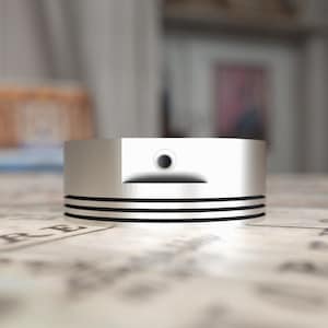 May include: A silver piston ring with three black bands around the circumference. The ring has a small hole in the center.