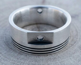 May include: A silver-toned metal ring with a brushed finish, featuring a flat rectangular section on the outer band with a small circular hole. The lower portion of the ring is decorated with three thin, parallel horizontal black engraved lines.