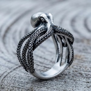 May include: A silver-toned ring shaped like an octopus with its tentacles wrapping around the band. The metal features a dark, oxidized finish in the recessed areas to highlight the detailed circular suckers along each of the octopus arms.