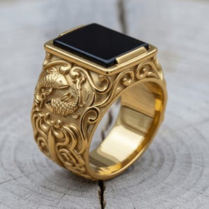 May include: A gold-toned ring featuring a rectangular black stone inlay on the top. The band is decorated with intricate relief carvings of two fish surrounded by swirling, wave-like patterns that extend around the entire outer surface.