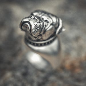 以下が含まれることがあります： ブルドッグの頭部が特徴的なシルバーリング。リングは磨き上げられた仕上げで、犬の顔の特徴と首輪を際立たせています。デザインは、ぼやけた質感の背景に配置されています。