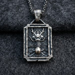 Puede incluir: Un collar con colgante de plata con un diseño detallado de dragón dentro de un marco rectangular. El dragón tiene detalles intrincados y está suspendido de una cadena de plata. El colgante está sobre un fondo gris oscuro.