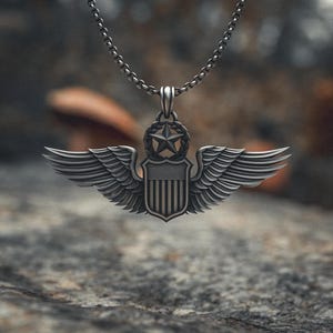 May include: A silver-colored pendant necklace featuring a detailed winged emblem with a star and shield design. The pendant hangs from a delicate chain, set against a blurred, natural background. The wings are spread wide, and the shield has vertical stripes.