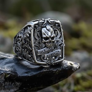 May include: A silver ring featuring a skull motif and the Latin inscription "Vita Brevis, Ars Longa". The ring has an intricate, detailed design and is displayed on a dark stone.