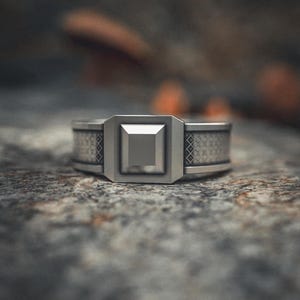 May include: A silver ring with a geometric design. The ring features a square center stone and a band with a repeating pattern. The ring is set on a textured surface.
