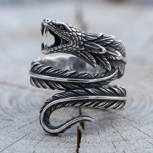 May include: A silver-toned metal ring shaped like a coiled snake with an open mouth showing fangs. The body of the snake features a detailed feather-like texture that wraps around the finger, creating a layered, spiraling design with dark oxidized accents.