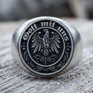 May include: A silver-toned metal ring featuring a circular face with an embossed eagle emblem at the center. The text "Gott mit uns" is engraved along the upper edge, with small cross symbols on the sides and oak leaf motifs at the bottom.