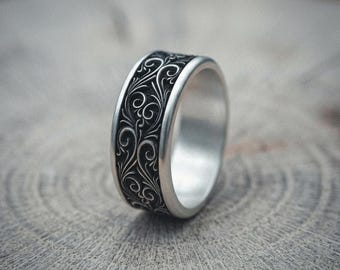 May include: A wide silver band ring featuring a recessed center channel with a dark, oxidized finish. The center band is decorated with intricate, raised silver scrollwork patterns that contrast against the black background of the metal.