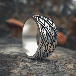 Puede incluir: Un anillo plateado con un detallado patrón en forma de pluma alrededor de la banda. El anillo se encuentra sobre un fondo natural borroso, resaltando el intrincado diseño y el brillo metálico.