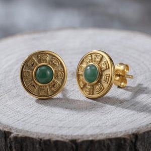 Op de afbeelding: Een paar goudkleurige oorknopjes met een cirkelvormig ontwerp met een reliëfpatroon rondom een centrale gladde groene steen. De oorbellen hebben een sluiting met een stift en een gepolijste metalen afwerking op de cirkelvormige voorzijde.