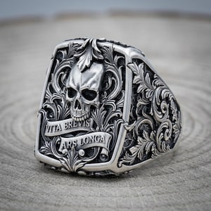 May include: A silver ring featuring a detailed skull centered within intricate scrollwork and acanthus leaf engravings. A banner across the bottom displays the engraved Latin text VITA BREVIS ARS LONGA. The metal has a polished finish.
