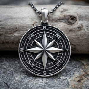 May include: A circular silver-toned pendant featuring a detailed compass rose design with cardinal and intercardinal directions marked as N, NE, E, SE, S, SW, W, and NW. The pendant has a textured dark background and hangs from a matching silver chain.