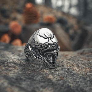 Könnte beinhalten: Ein silberner Ring mit einem detaillierten Design eines Venom-Charakters aus den Marvel-Comics. Der Ring ist aus Metall und hat eine strukturierte Oberfläche.