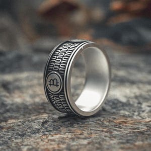 Puede incluir: Un anillo de plata con una banda negra que presenta un símbolo de Bitcoin y código binario. El anillo está sobre una superficie gris texturizada.
