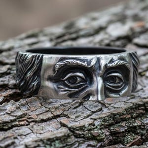 Könnte beinhalten: Ein silberner Ring mit detailliertem Gesichtsmotiv, einschließlich Augen und Haaren. Der Ring ist vor einem strukturierten, braunen Rindenhintergrund platziert. Das Design des Rings ist der Fokus, mit dunklem Innenraum.