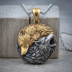 Op de afbeelding: Een ronde hanger met twee wolven in een yin-yang formatie, één in een gepolijste goudtint en de andere in een geoxideerde zilverkleur, hangend aan een zilverkleurige metalen ketting met een goudkleurig oogje aan de bovenkant van de hanger.