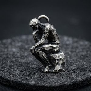 Puede incluir: Un colgante plateado con un diseño geométrico de una figura sentada, que recuerda a "El Pensador" de Rodin. El colgante tiene un lazo en la parte superior para una cadena. La figura está sentada sobre una base rocosa.