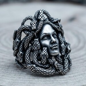 Könnte beinhalten: Ein silberfarbener Ring mit einem detaillierten skulpturalen Gesicht, das von mehreren ineinander verschlungenen Schlangen mit texturierten Schuppen umgeben ist. Das Metall hat ein poliertes Finish mit dunklen Vertiefungen zur Betonung.