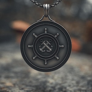 Könnte beinhalten: Silberfarbene Halskette mit einem runden Anhänger. Der Anhänger zeigt ein Steuerraddesign mit gekreuzten Ankern in der Mitte. Kette und Anhänger sind silberfarben. Der nautisch thematisierte Anhänger hängt an einer Schlaufe.