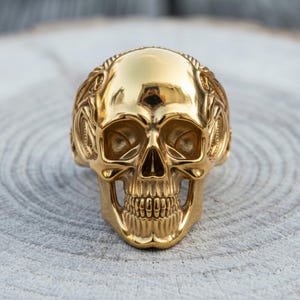 May include: A polished gold-tone ring featuring a detailed, three-dimensional human skull design. The skull has deep eye sockets, a defined nasal cavity, and visible teeth, with intricate, swirling decorative patterns carved along the sides.