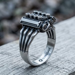 Könnte beinhalten: Ein silberfarbener Metallring mit einem detaillierten V8-Motorblock-Design auf der Oberseite. Die Ringschiene ist wie ein Abgaskrümmer geformt und enthält den eingravierten Text SLVR&SAGE mit einem kleinen Zahnradsymbol auf der Innenseite.