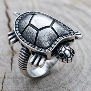 May include: A silver-toned ring shaped like a turtle with a textured shell, a border of small clear stones, and two black stones for the eyes. The band features a ribbed design, and the turtle's head, legs, and tail extend from the central shell.