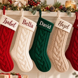 Op de afbeelding: Vijf gebreide kerstsokken met kabelpatroon hangen op een rij. De sokken zijn rood, wit, donkergroen en beige. Elke sok heeft een witte boord die is geborduurd met de namen Isabel, Bella, David, Hazel en Noah in rode of donkere tekst.