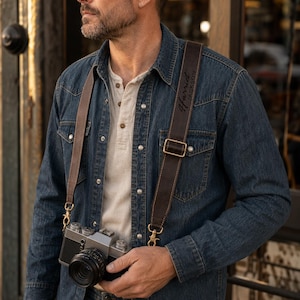 May include: A dark brown leather camera strap with visible stitching and brass-toned metal hardware, featuring the engraved name Harriet Jackson. The strap is attached to a silver and black camera, worn over a denim shirt and a light-colored henley shirt.