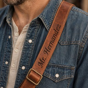 May include: A brown leather guitar strap featuring a distressed texture and visible stitching along the edges. The strap includes a brass-colored metal buckle for adjustment and is engraved with the custom text "Mr. Hernandez" in a dark script font.