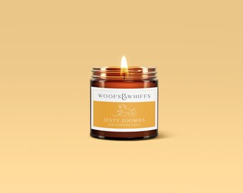 Op de afbeelding: Een brandende kaars in een bruine glazen pot met een zilveren deksel. Het etiket luidt "WOOFS & WHIFFS" boven "Zesty Zoomies" met een hondenafbeelding. De kaars staat op een lichtoranje achtergrond.