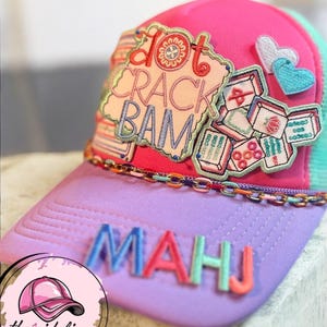May include: A pink, purple, and teal baseball cap with the word "MAHJ" in colorful letters. The cap features embroidered patches with the words "Dot Crack Bam" and images of mahjong tiles. A chain detail accents the brim.