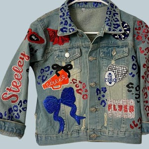 May include: Light blue denim jacket with various patches and embellishments. Features red and blue glitter accents, a leopard print collar, and the word "Steeley" on the sleeve. Patches include bows, a megaphone, and the words "Girl" and "Flyer".