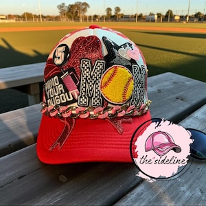 May include: A red baseball cap with a white top, featuring the word "MOM" in black and white letters, a yellow softball, and the text "MIND YOUR DUGOUT". The cap has a gold chain and pink accents.