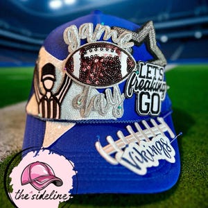 May include: A royal blue baseball cap with white and silver embellishments. The cap features the words "Game Day" and "Let's Freaking Go", along with a football and the word "Vikings". The cap has a pink logo that says "the sideline".
