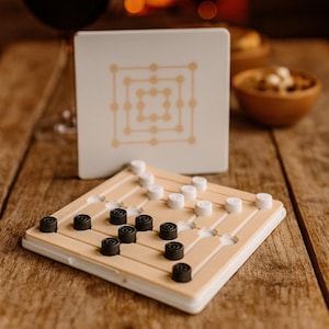 Könnte beinhalten: Ein Brettspiel mit schwarzen und weißen Spielsteinen, einer passenden Box und einem Glas Rotwein. Das Spiel befindet sich auf einem Holztisch mit einem verschwommenen Kamin im Hintergrund. Das Spielbrett ist beige mit einem Rastermuster.
