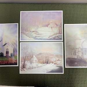 May include: Four framed art prints featuring winter landscape scenes with churches. The artwork has a soft, pastel color palette with snow-covered fields, trees, and buildings. The prints are arranged on a green textured surface.