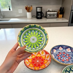 Puede incluir: Un conjunto de pequeños cuencos de cerámica decorativos con coloridos patrones geométricos. Los cuencos presentan diseños en verde, azul, rojo y amarillo, con un borde dorado. Los cuencos se exhiben sobre una encimera blanca.