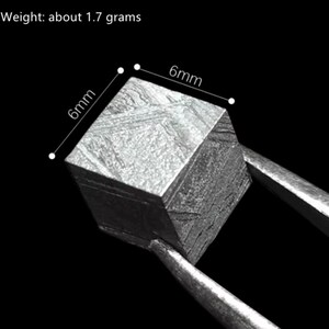 May include: A small, square, metallic object, approximately 6mm on each side, is held by tweezers. The object has a textured surface and weighs about 1.7 grams. The background is black.