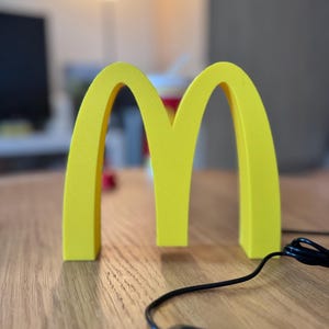 May include: A yellow, illuminated McDonald's logo, featuring the iconic golden arches. The logo is on a wooden surface, with a black cord and control panel visible. The background is blurred, suggesting an indoor setting.