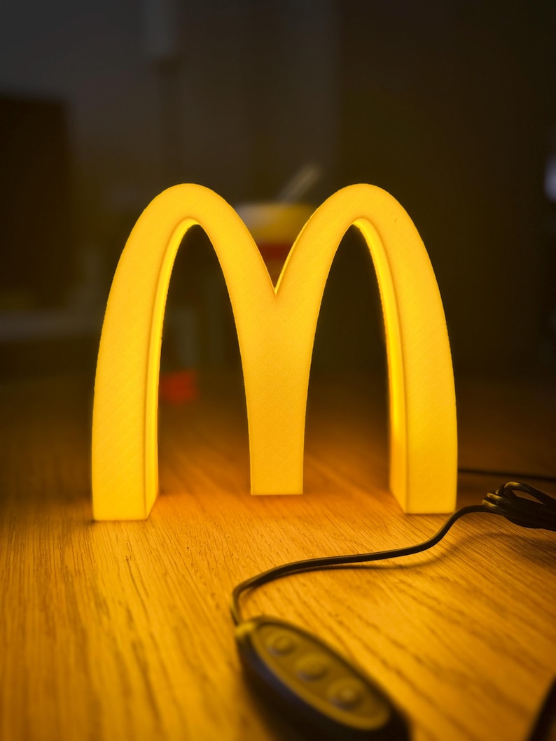 May include: A glowing yellow McDonald's logo, the iconic golden arches, illuminated against a dark background. The light is warm, and a black cord with a control is visible. The logo is on a wooden surface.