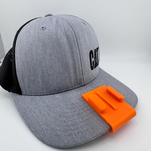 May include: Gray baseball cap with a black mesh back and the word "CAT" in black. An orange rectangular mount is attached to the brim, designed for a camera or accessory.