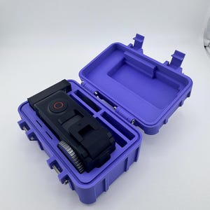 May include: A purple, open, rectangular case with a black camera inside. The case has a hinged lid and secure latches. The camera has a red button and a silver wheel. The case is designed to protect the camera.