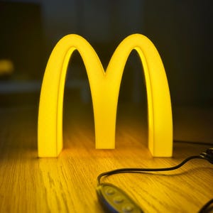 May include: A glowing yellow McDonald's logo, the iconic golden arches, illuminated against a dark background. The light is on a wooden surface, with a black cord and a remote control.